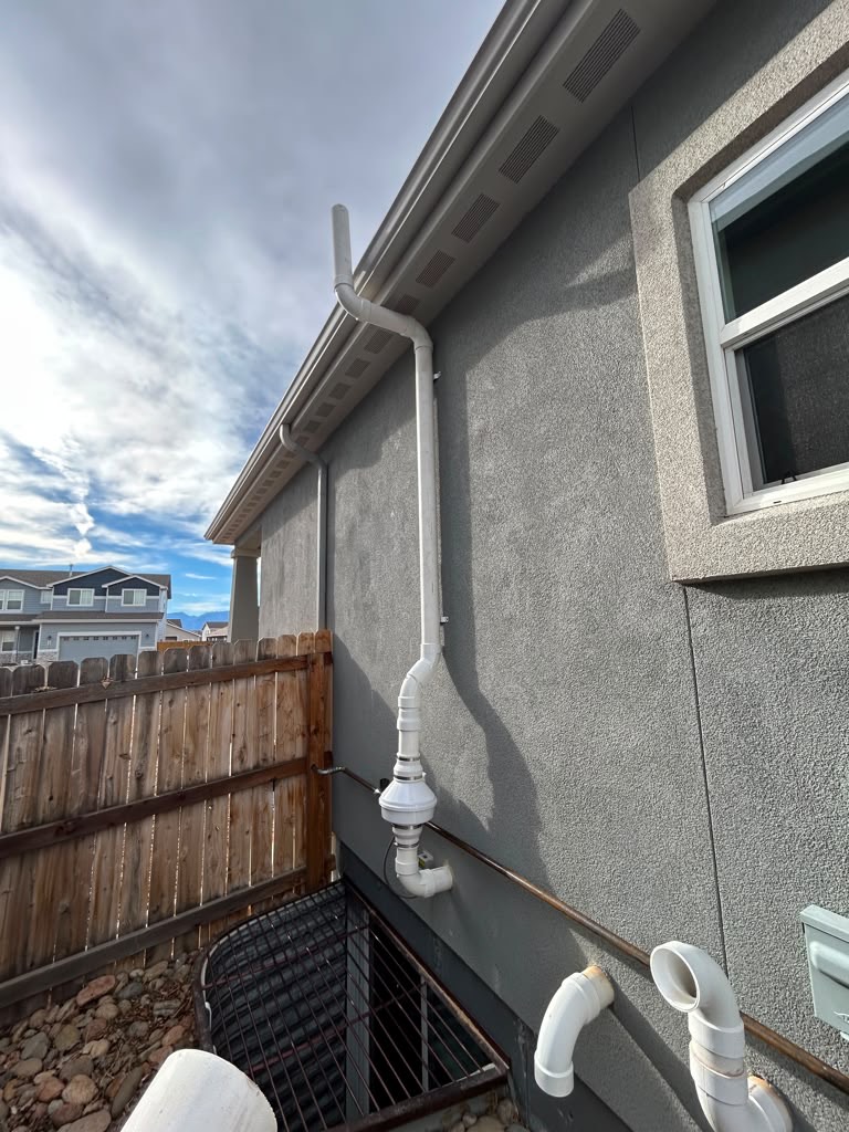 Exterior radon mitigation system on a stucco home, vent pipe running to the roofline with the inline fan above the window well